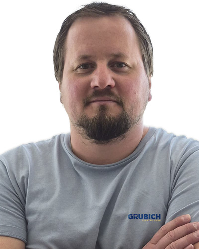 Mann mit verschränkten Armen trägt graues T-Shirt mit blauem Schriftzug 'GRUBICH' vor hellblauem Hintergrund